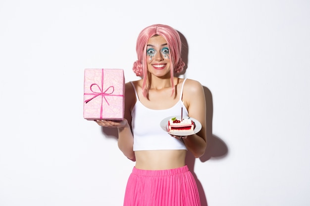 Ritratto della ragazza di compleanno eccitata che celebra la sua vacanza, che tiene il regalo di b-day e la torta, sorridendo felice, in piedi.