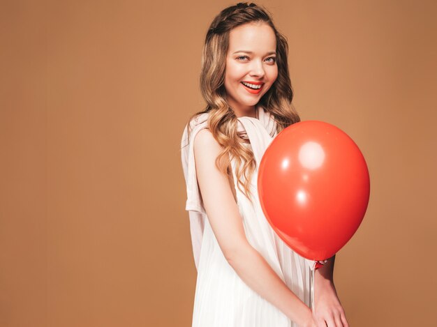 Ritratto della ragazza emozionante che posa in vestito bianco da estate alla moda. Donna sorridente con la posa rossa del pallone. Modello pronto per la festa