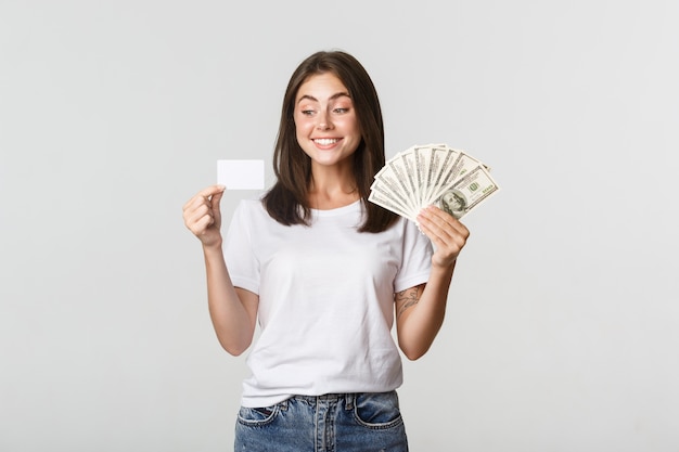 Ritratto della ragazza sorridente eccitata che tiene soldi e carta di credito, bianco.