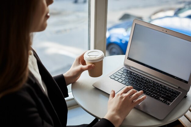 Ritratto della studentessa moderna che si siede nel caffè mentre beve il caffè e controllando la posta con il computer portatile