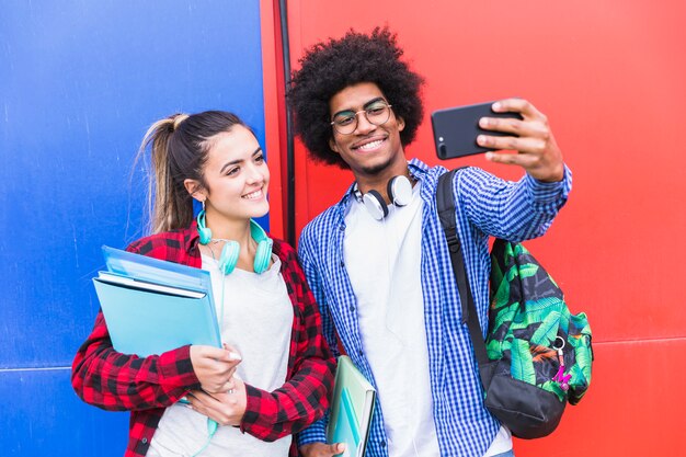 Ritratto delle coppie adolescenti sorridenti che prendono insieme selfie sul cellulare contro la parete colorata