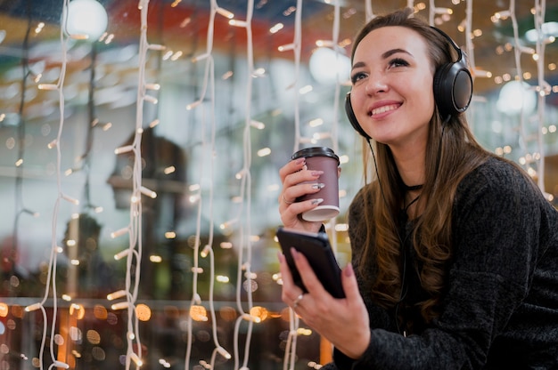 Ritratto delle cuffie da portare sorridenti della donna che tengono tazza e telefono vicino alle luci di natale