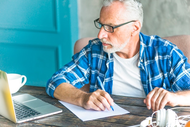 Ritratto delle note di scrittura di un uomo senior facendo uso del computer portatile sul tavolo