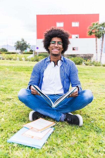 Ritratto dello studente maschio africano sorridente che si siede sul libro della tenuta dell&#39;erba verde nella mano