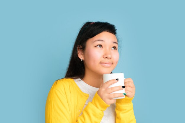 Ritratto di adolescente asiatico isolato su sfondo blu studio. Bello modello femminile del brunette con i capelli lunghi. Concetto di emozioni umane, espressione facciale, vendite, annuncio. Bere caffè o tè.