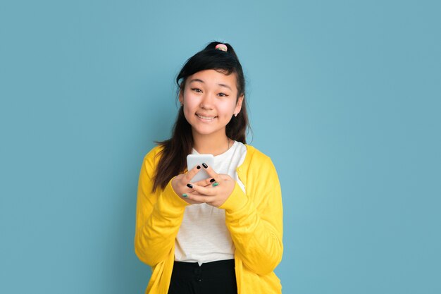 Ritratto di adolescente asiatico isolato su sfondo blu studio. Bello modello femminile del brunette con i capelli lunghi. Concetto di emozioni umane, espressione facciale, vendite, annuncio. Usando il telefono, sorridendo.