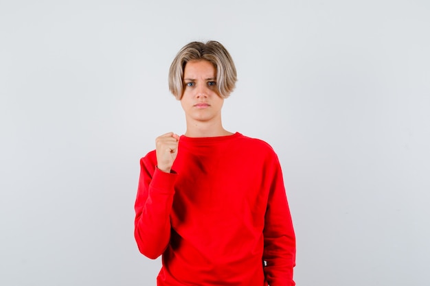 Ritratto di adolescente maschio biondo avvertimento con pugno in maglione rosso e guardando arrabbiato vista frontale