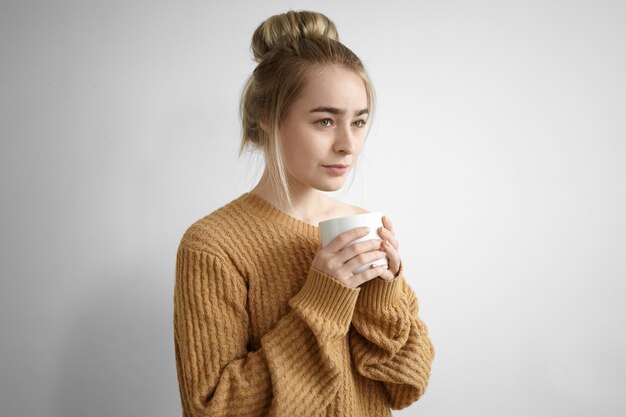 Ritratto di adorabile affascinante giovane donna con pettinatura disordinata che tiene grande tazza, avendo il caffè del mattino fresco a casa