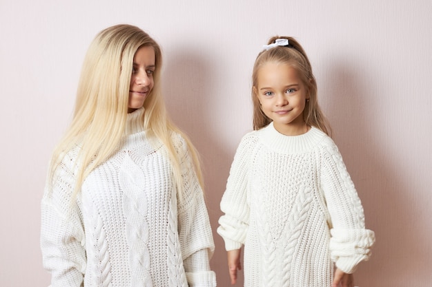 Ritratto di adorabile bambina carina che indossa maglione lavorato a maglia e nastro nei capelli sorridente mentre trascorre del tempo piacevole con la sua premurosa madre amorevole che sta guardando la figlia con amore e tenerezza