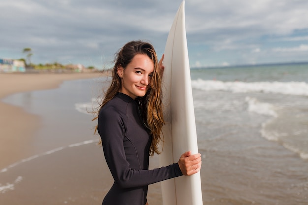 Ritratto di adorabile giovane donna amichevole con lunghi capelli castano chiaro e grandi occhi si leva in piedi sulla riva dell'oceano con emozioni vere felici e si prepara per la lezione di surf