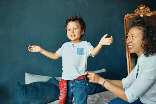 Ritratto di adorabile ragazzino con i capelli ricci in piedi sul letto, esprimendo emozioni positive, giovane madre guardando il figlio con orgoglio e affetto.