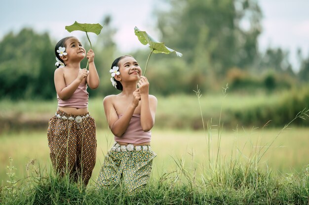 Ritratto di adorabile sorella e giovane sorella in abito tradizionale tailandese e mettere un fiore bianco sul suo orecchio, guardando la foglia di loto in mano e sorridendo con felice sul campo di riso, copia spazio