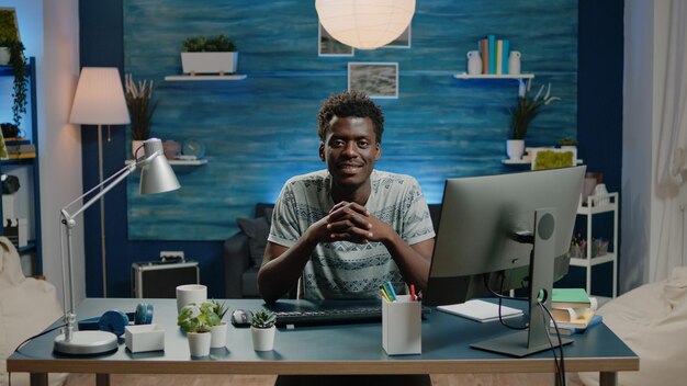 Ritratto di adulto nero seduto alla scrivania con computer e notebook. Uomo di etnia africana che guarda la macchina fotografica e sorride mentre ha un gadget per il lavoro aziendale remoto online.