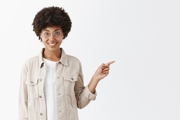 Ritratto di affascinante afroamericano in occhiali e camicia con acconciatura afro, che punta a destra con il dito indice e sorridente