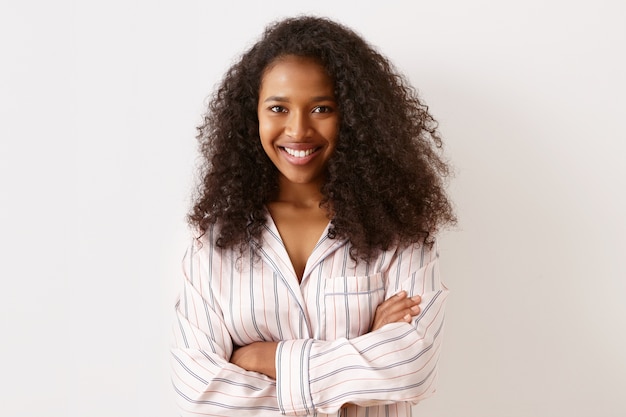 Ritratto di affascinante attraente giovane donna afro-americana con voluminoso taglio di capelli afro con un ampio sorriso fiducioso, mantenendo le braccia incrociate sul petto, indossa una camicia da notte a strisce