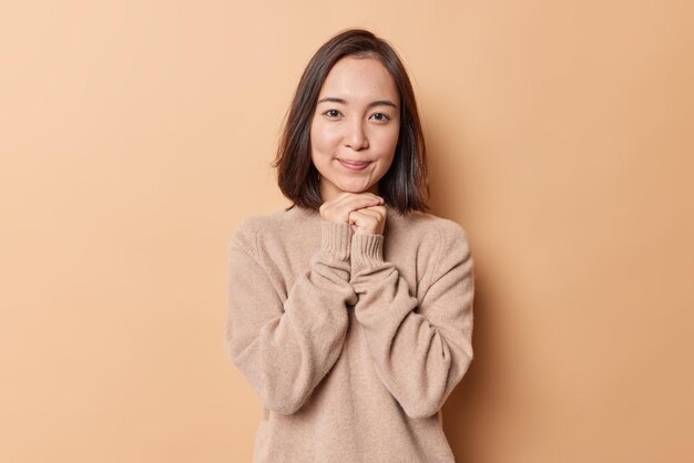 Ritratto di affascinante bruna donna asiatica tiene le mani sotto il mento sorride delicatamente indossa un maglione caldo morbido isolato su sfondo beige. La modella piuttosto giovane con la pelle pulita si sente soddisfatta