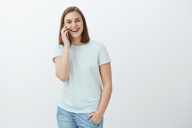 Ritratto di affascinante donna felice e intrattenuta che tiene smartphone vicino all'orecchio chiamando e parlando divertito sopra il muro bianco