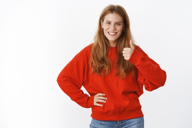 Ritratto di affascinante donna sincera carina in maglione rosso caldo che mostra pollice in su gesto tenendo la mano sulla vita e sorridente dando risposta positiva, gradimento idea