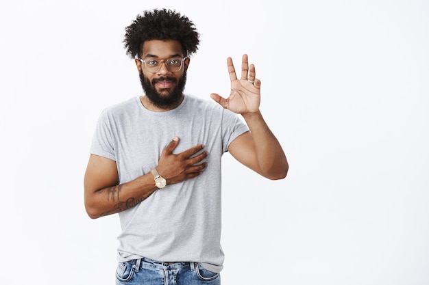 Ritratto di affascinante fiducioso, sorridente uomo afroamericano che dà promessa alzando la mano e tenendo il braccio sul cuore come dando giuramento o impegno, giurando di non mentire sul muro grigio