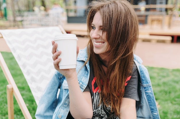 Ritratto di affascinante giovane donna con lunghi capelli scuri vestita in giacca di jeans è seduta nel parco con una tazza di caffè e guarda da parte con un grande sorriso. Buona giornata di sole. Umore rilassato.