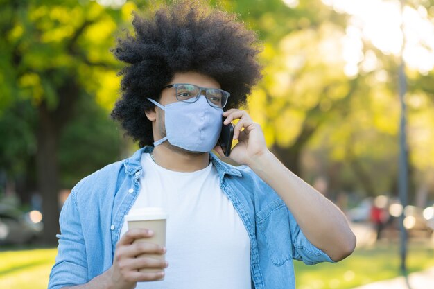 Ritratto di afro latino uomo che indossa la maschera per il viso e parla al telefono mentre in piedi all'aperto sulla strada