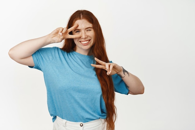 Ritratto di allegra studentessa rossa ragazza con lunga acconciatura naturale che mostra pace vsigns vicino agli occhi e strizzando l'occhio carino alla telecamera godersi l'evento divertendosi sfondo bianco