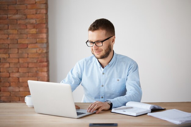 Ritratto di allegro bello con la barba lunga manager di società maschile in bicchieri e abbigliamento casual seduto al tavolo in ufficio, sorridendo dolcemente, guardando il monitor del computer portatile, essendo felice di fare il lavoro preferito