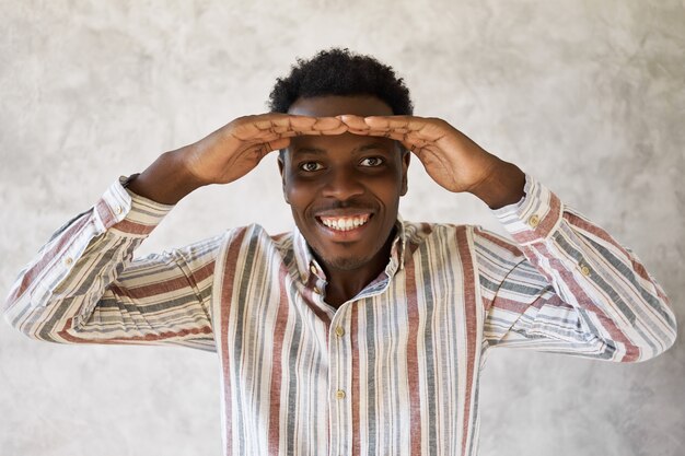 Ritratto di allegro felice giovane ragazzo afroamericano in camicia casual che esamina la distanza tenendo entrambe le mani sulla fronte e sorridendo ampiamente, essendo entusiasta del futuro luminoso. Linguaggio del corpo