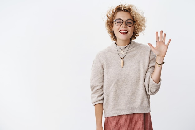 Ritratto di amichevole dall'aspetto estroverso e rilassato spensierato attraente ragazza hipster con occhiali e maglione alzando il palmo per salutare i nuovi membri del gruppo sorridendo gioiosamente