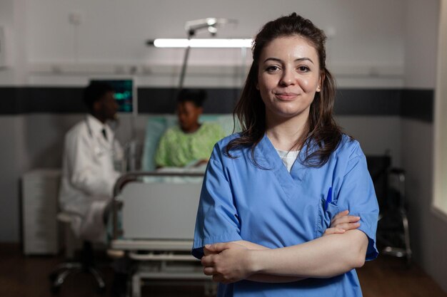 Ritratto di assistente medico specialista in piedi nel reparto ospedaliero durante la consultazione medica. In background medico consulto paziente malato discutendo il trattamento sanitario. Servizi di medicina