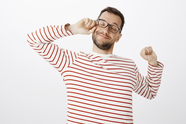 Ritratto di assonnato stanco contento ragazzo con gli occhiali, strofinando gli occhi e sorridendo ampiamente mentre si allunga dall'esaurimento, volendo dormire dopo una lunga giornata di lavoro