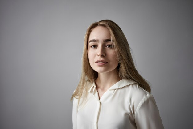 Ritratto di attraente alla moda giovane bionda signora caucasica con le lentiggini, pettinatura sciolta e nessun trucco in posa in studio indossando una camicia bianca al muro grigio copyspace, guardando la fotocamera