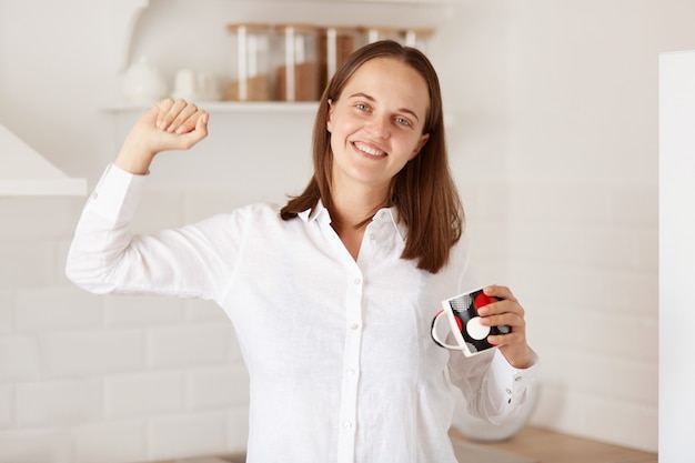 Ritratto di attraente donna dai capelli scuri che indossa una camicia casual bianca, guardando la telecamera con un sorriso affascinante e allungando le braccia, godendosi il caffè caldo dopo il risveglio.