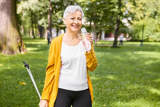 Ritratto di attraente donna senior in buona salute con capelli grigi pixie riposando mentre si cammina nel parco utilizzando i poli scandinavi nordici, tenendo la bottiglia, acqua potabile, sentendosi pieni di energia, sorridente