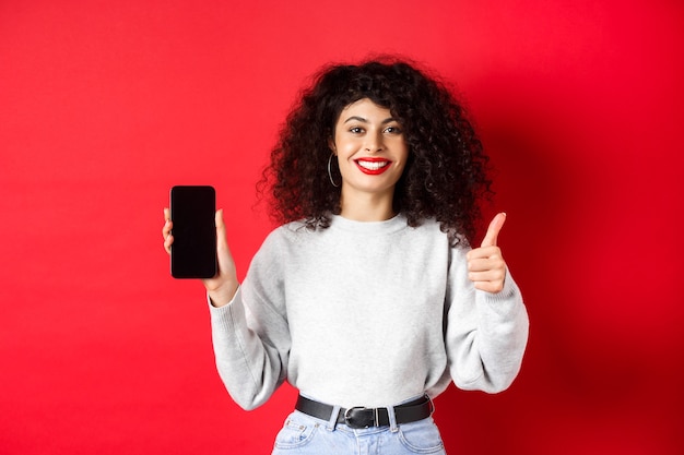 Ritratto di attraente donna sorridente con i capelli ricci, che mostra lo schermo del telefono cellulare vuoto e il pollice in su, raccomandando promo online, in piedi su sfondo rosso