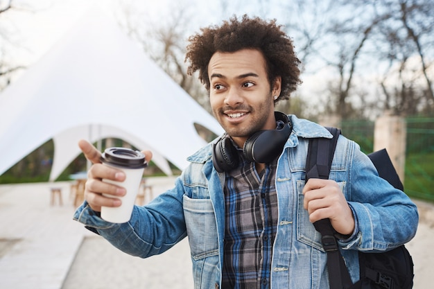 Ritratto di attraente giovane afro-americano con acconciatura afro che tiene zaino e caffè, indossando abiti alla moda e cuffie sopra il collo, indicando da qualche parte