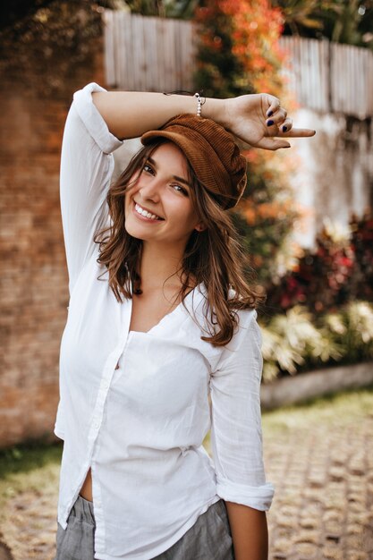 Ritratto di attraente giovane donna dagli occhi marroni in camicia di cotone oversize e cappello di velluto a coste con il sorriso che guarda l'obbiettivo in cortile.