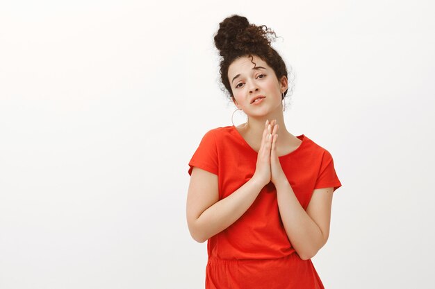 Ritratto di avida donna carina con i capelli ricci in abito rosso, tenendo le mani in preghiera con i palmi insieme sul petto, implorando favore o aiuto