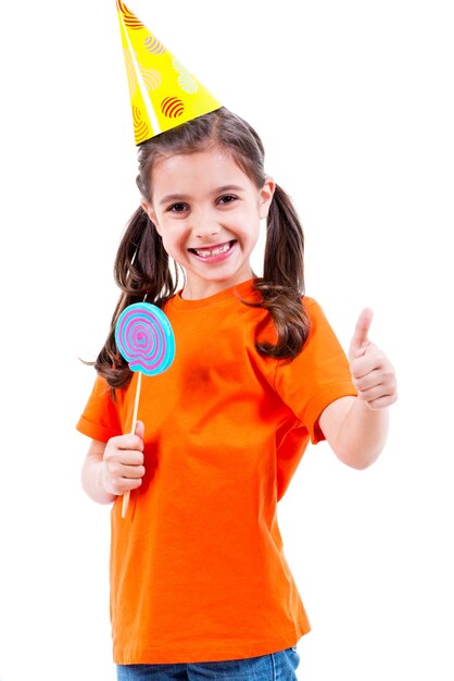 Ritratto di bambina carina in maglietta arancione e cappello da festa con caramelle colorate che mostra i pollici aumenta il gesto - isolato su bianco