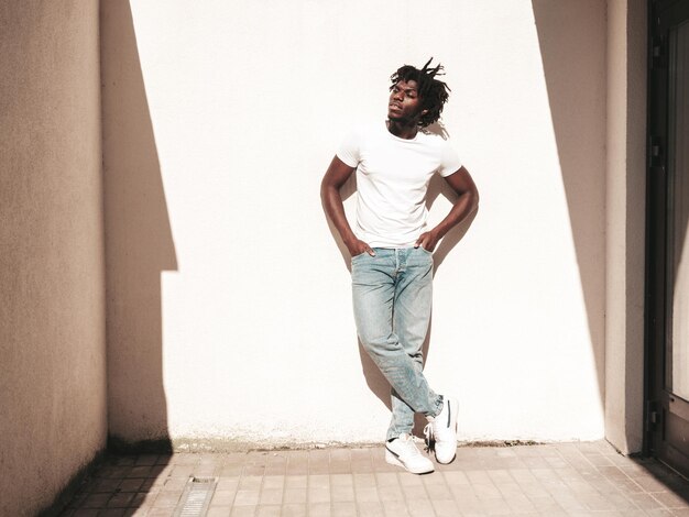 Ritratto di bel modello hipsterUnshaven africano uomo vestito con maglietta bianca estiva e jeans Moda maschile con acconciatura di dreadlocks in posa vicino al muro bianco in strada