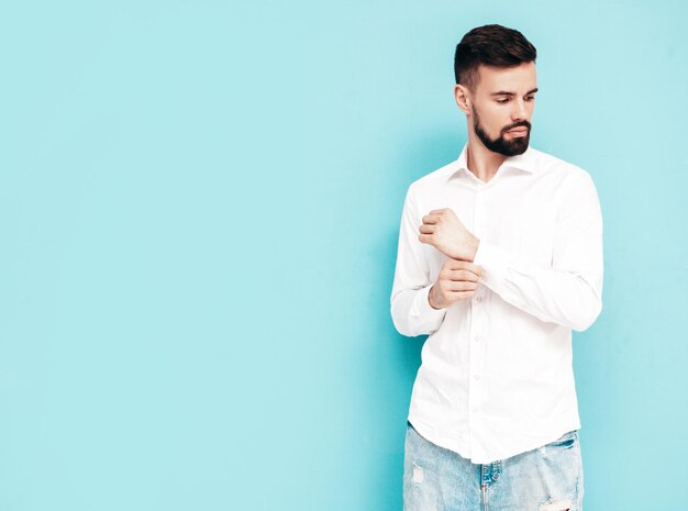 Ritratto di bel modello sorridente Uomo alla moda sexy vestito in camicia bianca e jeans Moda maschio hipster in posa vicino al muro blu in studio isolato