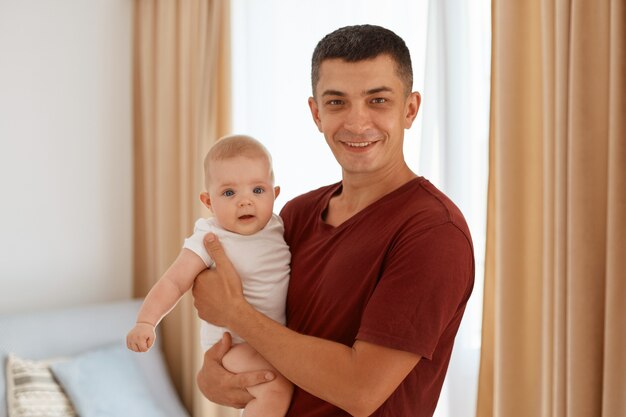 Ritratto di bel padre che indossa una maglietta bordeaux con un'affascinante figlia neonata in mano, che guarda l'obbiettivo, in piedi in un accogliente soggiorno con finestra sullo sfondo.