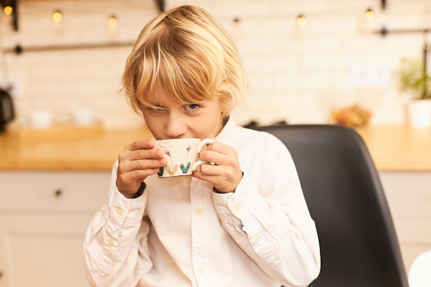 Ritratto di bel ragazzo gioioso con capelli biondi, bere il tè mentre si fa colazione prima della scuola, tenendo la tazza e sorridente con utensili e ghirlanda sul bancone della cucina