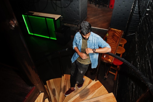 Ritratto di bel successo barbuto del sud asiatico giovane libero professionista indiano in camicia di jeans blu in piedi nel night club sulle scale