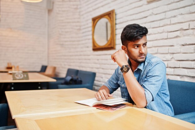 Ritratto di bel successo barbuto del sud asiatico giovane libero professionista indiano in camicia di jeans blu seduto in caffè e menu di lettura