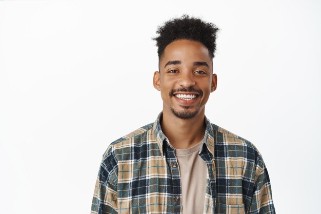 Ritratto di bell'uomo afroamericano con baffi, denti bianchi sorridenti, che sembra felice e sicuro di sé, in piedi in camicia a quadri su sfondo bianco