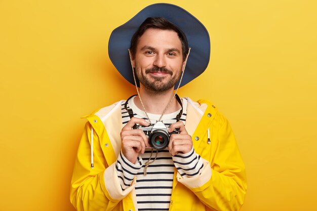 Ritratto di bell'uomo con la barba lunga tiene la fotocamera retrò, scatta foto di qualcosa, indossa cappello, maglione a righe e impermeabile, isolato sopra il muro giallo