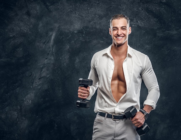 Ritratto di bell'uomo muscoloso con manubri in camicia bianca in studio fotografico scuro.