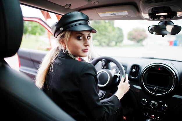 Ritratto di bella bionda modello di donna di moda sexy in berretto e in tutto nero con trucco luminoso sedersi e guidare un'auto da città rossa
