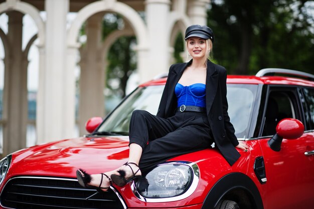 Ritratto di bella bionda modello di donna di moda sexy in berretto e in tutto nero con trucco luminoso sedersi sul cofano della city car rossa